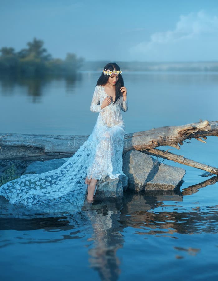 Eine Nymphe mit dem langen dunklen Haar in einem weißen Weinlesekleid sitzt auf einem Klotz über dem Fluss im Haar ein Kranz von stockbild Eine Nymphe mit dem langen dunklen Haar in einem weißen Weinlesekleid sitzt auf einem Klotz über dem Fluss im Haar ein Kranz von stockbild