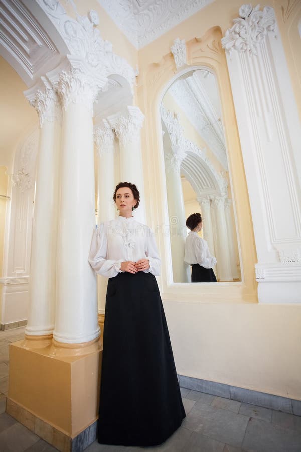 Eine respektable Frau in uralten ukrainischen Kleidung in einem schwarzen langen Rock und einer weißen Bluse. Eine respektable Frau in ukrainischer Kleidung in einem schwarzen langen Rock und eine weiße Bluse steht in den Räumlichkeiten eines schicken barocken Anwesen mit großen Säulen und einem Spiegel