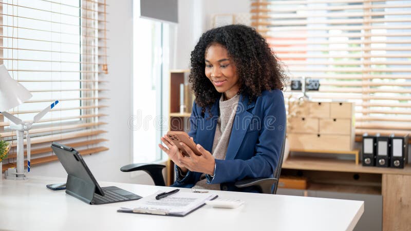 Eine schwarze Immobilienmaklerin überprüft ihre Nachrichten auf ihrem Smartphone an ihrem Schreibtisch stockfotografie