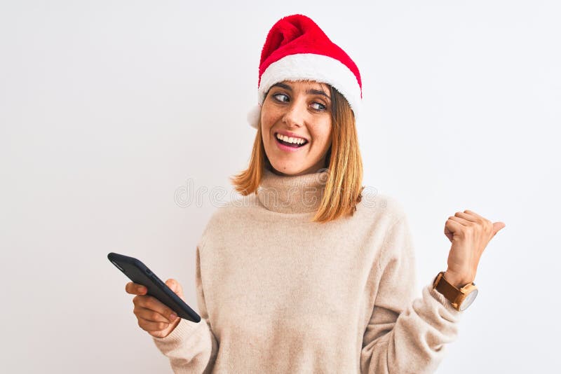 Eine schöne Rothoutiquin mit Weihnachtsmütze und Smartphone-Zeige und mit Daumen bis zur Seite mit fröhlichem Gesicht lizenzfreie stockfotografie