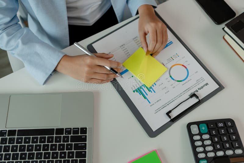 Eine Unternehmerin arbeitet in ihrem Büro und schreibt eine Notiz, die eine weibliche Mitarbeiterin schreibt, um eine kurze Zusammenfassung eines Berichts zu erstellen, damit sie während der Arbeitszeit nicht vergessen wird, am Arbeitsplatz zu arbeiten