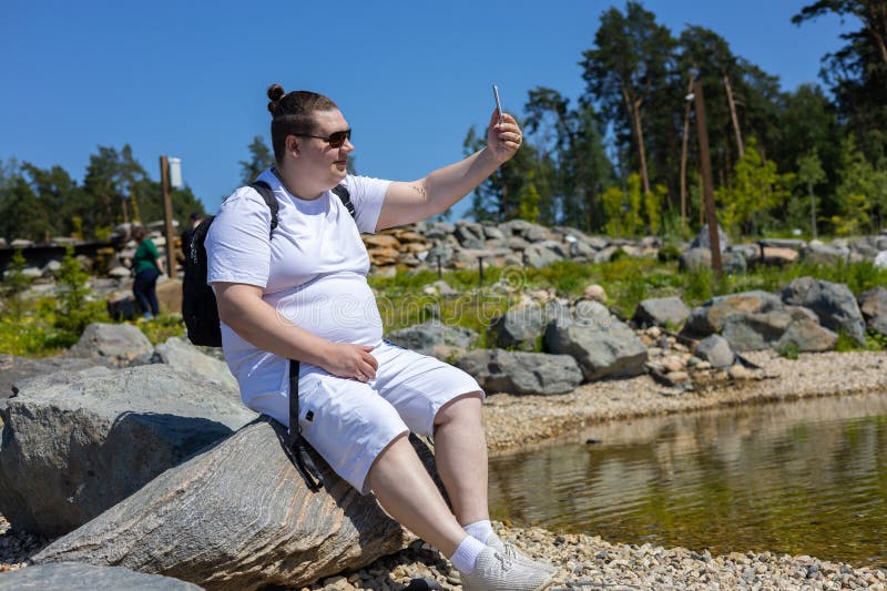 An einem sommerlichen sonnigen Tag sitzt ein Mann auf einem Stein am See und nimmt ein Selfie am Telefon. ein Mann für stockbild