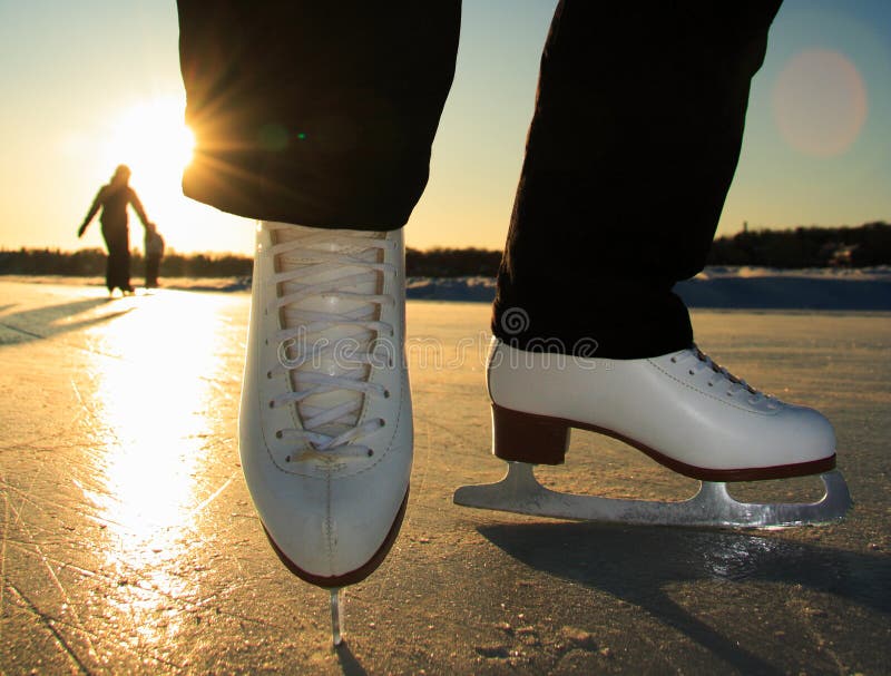 Ice skating. Ice skates in action closeup outdoors. Classic figure ice skates on frozen lake outdoors in evening light, Mom and daughter silhouette in the background. Lac Beauport, Quebec City, Canada. Ice skating. Ice skates in action closeup outdoors. Classic figure ice skates on frozen lake outdoors in evening light, Mom and daughter silhouette in the background. Lac Beauport, Quebec City, Canada.