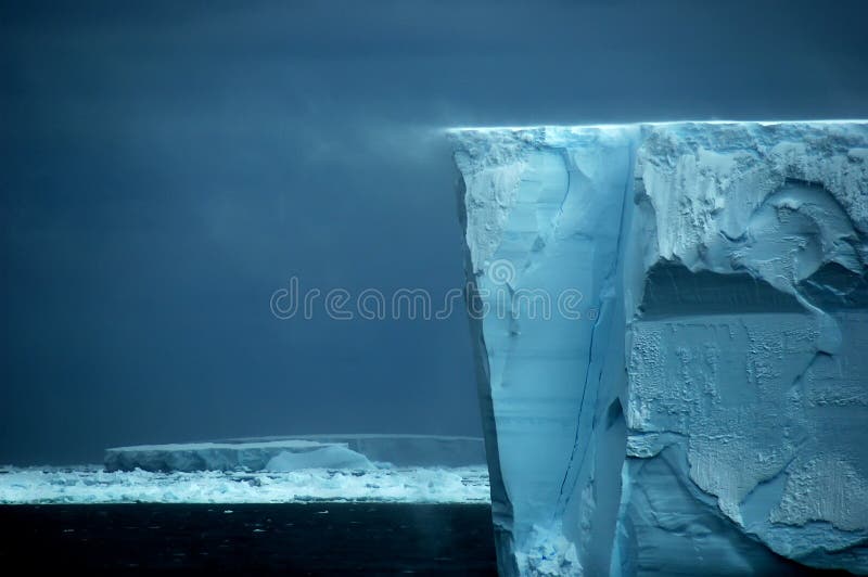 The Antarctic ice shelf edge at snow drift. The ice wall shows a nice relief pattern. In the background a large table iceberg. Picture was taken during a 3-month Antarctic research expedition near the Peninsula. The Antarctic ice shelf edge at snow drift. The ice wall shows a nice relief pattern. In the background a large table iceberg. Picture was taken during a 3-month Antarctic research expedition near the Peninsula.