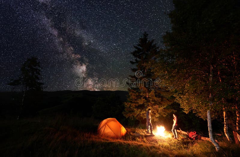 El acampar de la noche Fuego que hace una pausa del individuo y de la muchacha cerca del bosque y de las tiendas debajo del cielo imagenes de archivo El acampar de la noche Fuego que hace una pausa del individuo y de la muchacha cerca del bosque y de las tiendas debajo del cielo imagenes de archivo