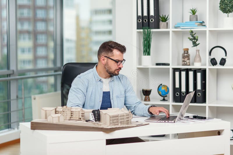 El arquitecto comprometido trabaja en la oficina de diseño con el ordenador portátil y se examinan los detalles en el modelo de la fotografía de archivo libre de regalías