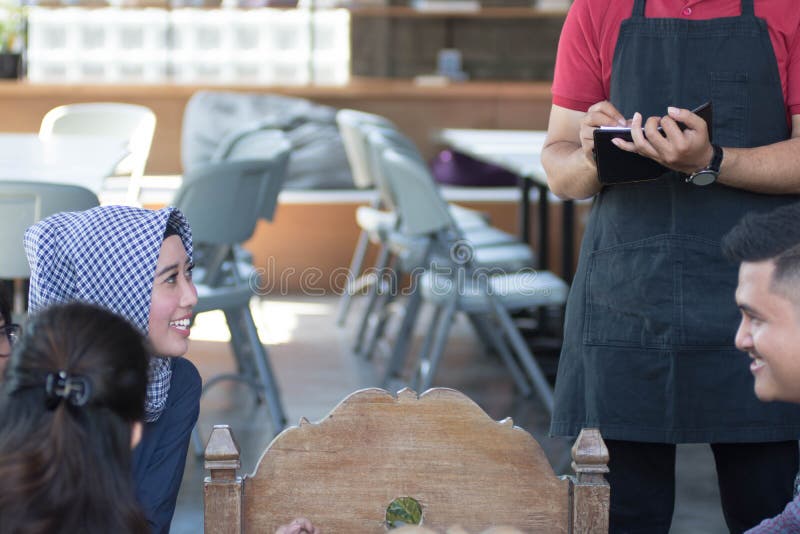 El camarero de sexo masculino asiático escribe órdenes de costumers en el café en fondo imagenes de archivo El camarero de sexo masculino asiático escribe órdenes de costumers en el café en fondo imagenes de archivo