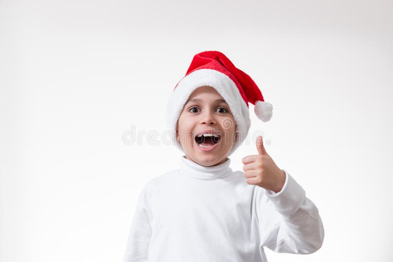 El chico en el sombrero rojo de santa ríe en voz alta y muestra su dedo pulgar. Concepto de Navidad imagenes de archivo