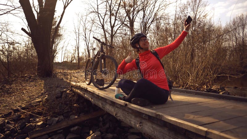 El ciclista cauc?sico feliz de la mujer hace una foto de s? misma un selfie en el tel?fono mientras que asiste en el puente sobre fotografía de archivo libre de regalías El ciclista cauc?sico feliz de la mujer hace una foto de s? misma un selfie en el tel?fono mientras que asiste en el puente sobre fotografía de archivo libre de regalías