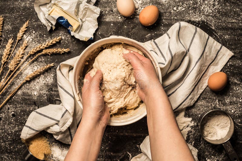 Baking - cook`s hands kneading the raw dough pastry in a bowl. Wheat, butter, eggs, flour and sugar around. Kitchen scenery from above top view, flat lay on black chalkboard background. Baking - cook`s hands kneading the raw dough pastry in a bowl. Wheat, butter, eggs, flour and sugar around. Kitchen scenery from above top view, flat lay on black chalkboard background.