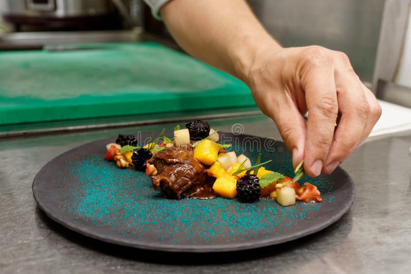 El cocinero está adornando la ternera guisada con las bayas, las hierbas y la salsa de la fruta, cocina comercial fotografía de archivo El cocinero está adornando la ternera guisada con las bayas, las hierbas y la salsa de la fruta, cocina comercial fotografía de archivo
