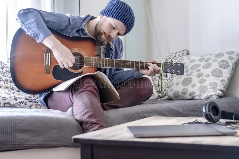 El compositor enfocado juega música escrita en una guitarra acústica mientras que mira un cuaderno y con un lápiz en su boca, sen