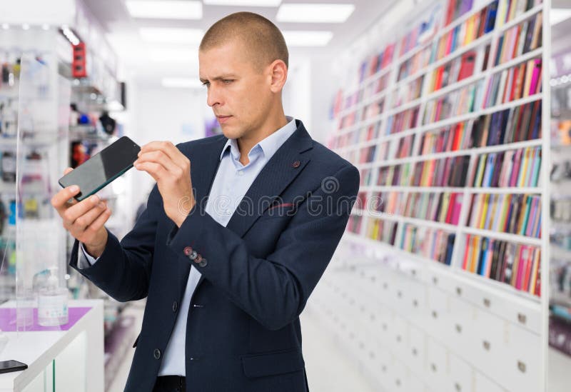 Smiling elegant young man standing in tech store with new smart phone in his hands. Smiling elegant young man standing in tech store with new smart phone in his hands