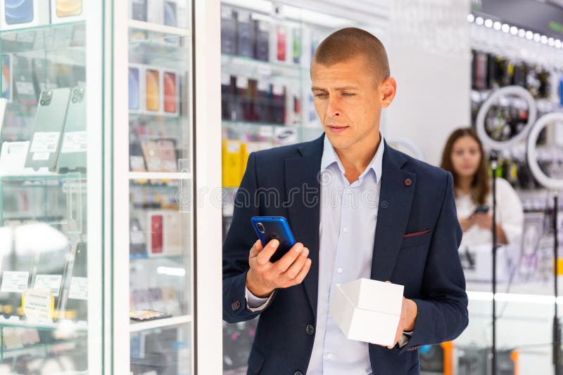 Smiling elegant young man standing in tech store with new smart phone in his hands. Smiling elegant young man standing in tech store with new smart phone in his hands