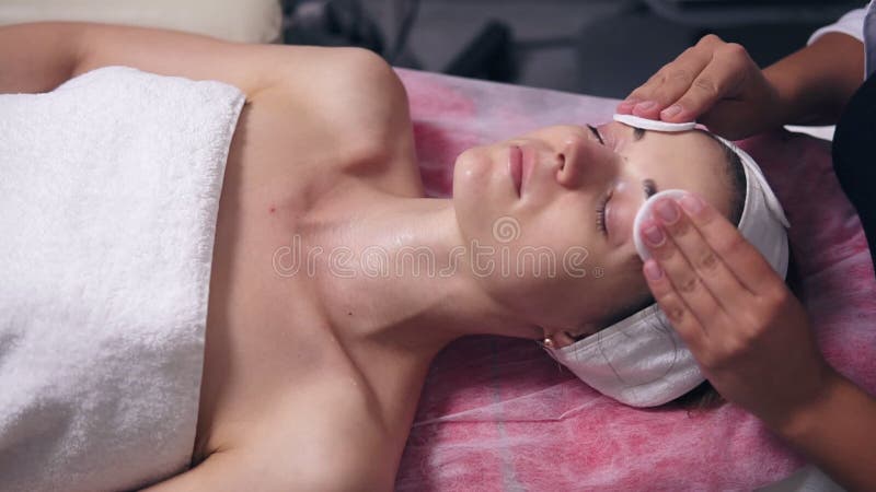 El cosmetologist profesional está entonando la cara del ` s de la mujer usando esponja del algodón La mujer joven está mintiendo