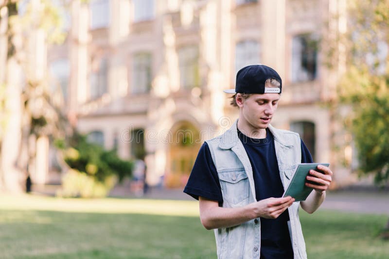 The young student stands on the background of the university and reads the task from his notebook. The young student stands on the background of the university and reads the task from his notebook.