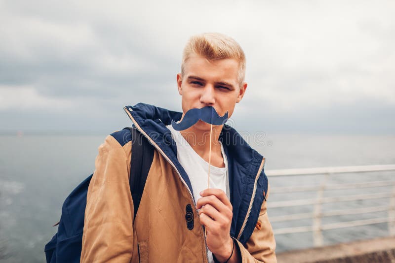El estudiante universitario con la mochila que sostiene la cabina de la foto apoya el bigote por el mar Individuo que se divierte foto de archivo libre de regalías El estudiante universitario con la mochila que sostiene la cabina de la foto apoya el bigote por el mar Individuo que se divierte foto de archivo libre de regalías