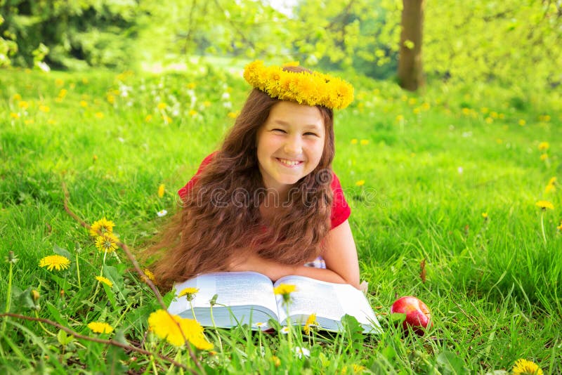 Summer portrait, merry child among the dandelions. Smiling girl with crown of dandelions flowers lies on the green grass on the lawn. Child reads thick book with a red apple. Soon back to school!. Summer portrait, merry child among the dandelions. Smiling girl with crown of dandelions flowers lies on the green grass on the lawn. Child reads thick book with a red apple. Soon back to school!