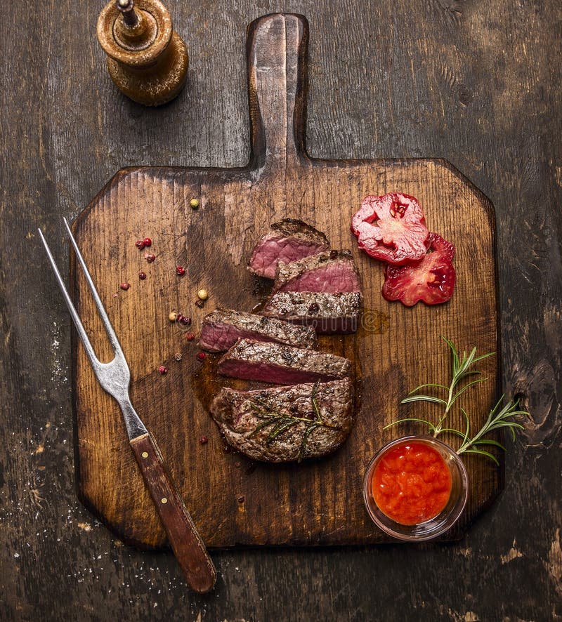 El filete asado del ribeye cortó el ‹del †del ‹del †en una tabla de cortar con una bifurcación, una salsa roja, pimientas y t imagen de archivo libre de regalías El filete asado del ribeye cortó el ‹del †del ‹del †en una tabla de cortar con una bifurcación, una salsa roja, pimientas y t imagen de archivo libre de regalías