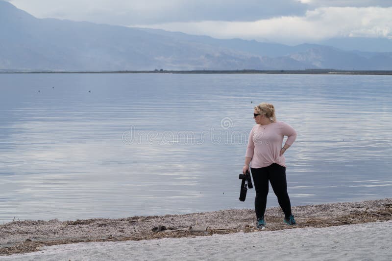 El fotógrafo de sexo femenino se coloca en la línea de la playa del mar de Salton, con su cámara, mirando fijamente apagado en fotografía de archivo El fotógrafo de sexo femenino se coloca en la línea de la playa del mar de Salton, con su cámara, mirando fijamente apagado en fotografía de archivo