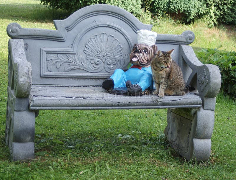 El gato bonito se sienta al lado de la figura de un dogo inglés en el banco del jardín imágenes de archivo libres de regalías El gato bonito se sienta al lado de la figura de un dogo inglés en el banco del jardín imágenes de archivo libres de regalías