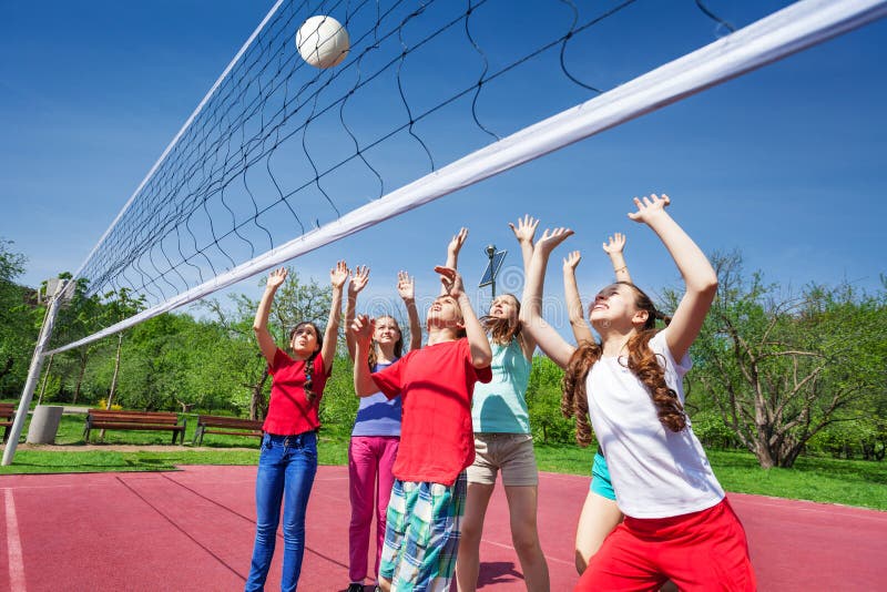 Group of teens with arms up play volleyball near the net on the court during sunny summer day outside. Group of teens with arms up play volleyball near the net on the court during sunny summer day outside