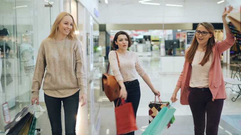 El grupo de muchachas felices se está divirtiendo en centro comercial que caminan con las bolsas de papel, riendo y bailando seña