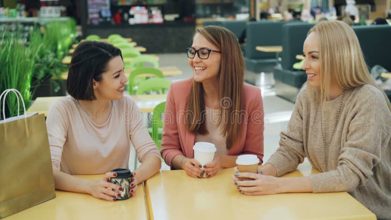 El grupo de señoras jovenes felices es de charla y de risa de la sentada de la tabla en café del centro comercial con las bebidas