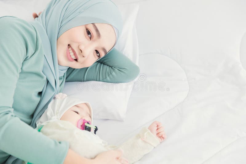 El hijab musulmán se queda con su bebé en cama blanca sonriendo en casa, la madre cuida de un bebé fotografía de archivo libre de regalías