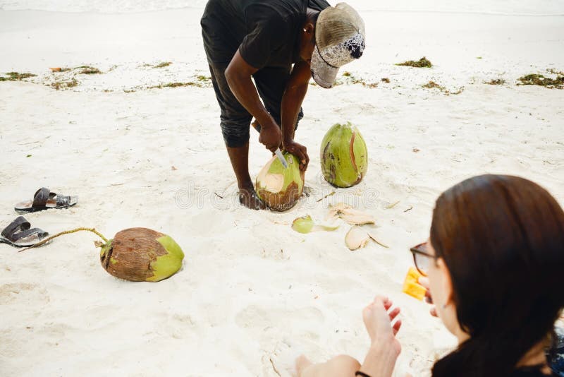 El hombre africano pela el coco para el turista femenino en la playa foto de archivo libre de regalías El hombre africano pela el coco para el turista femenino en la playa foto de archivo libre de regalías