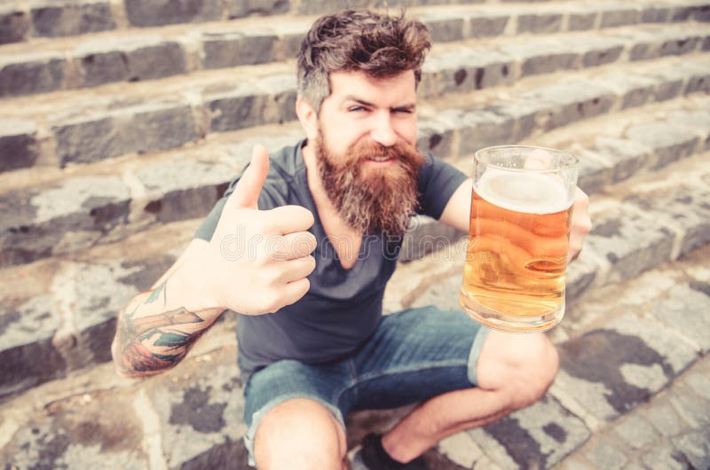 El hombre con la barba y el bigote sostiene el vidrio con la cerveza y muestra el pulgar para arriba, el fondo de piedra de las e imágenes de archivo libres de regalías El hombre con la barba y el bigote sostiene el vidrio con la cerveza y muestra el pulgar para arriba, el fondo de piedra de las e imágenes de archivo libres de regalías