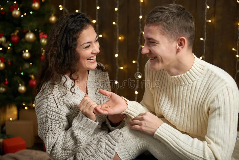 El hombre da a muchacha un anillo de compromiso, pares en luces de la Navidad y la decoración, vestida en blanco, árbol de abeto imágenes de archivo libres de regalías El hombre da a muchacha un anillo de compromiso, pares en luces de la Navidad y la decoración, vestida en blanco, árbol de abeto imágenes de archivo libres de regalías