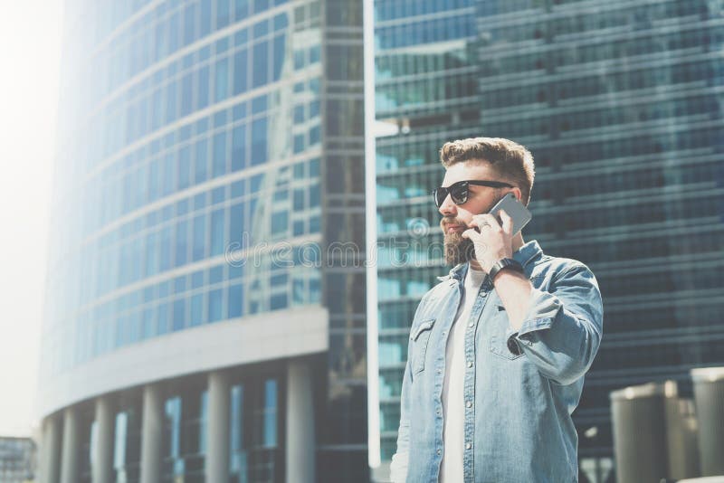 El hombre de negocios barbudo joven en gafas de sol y camisa se coloca en la calle de la ciudad y habla en su teléfono celular Dí foto de archivo libre de regalías El hombre de negocios barbudo joven en gafas de sol y camisa se coloca en la calle de la ciudad y habla en su teléfono celular Dí foto de archivo libre de regalías