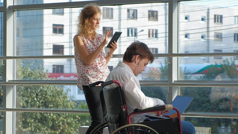 El hombre de negocios discapacitado en una silla de ruedas en una ventana con un ordenador portátil cerca de la mujer utiliza una metrajes