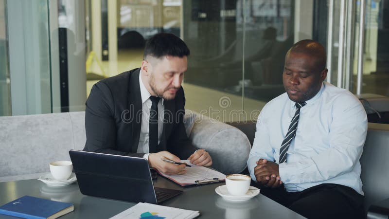 El hombre de negocios enojado en traje de negocios negro critica seriamente a su empleado afroamericano durante la reunión en caf