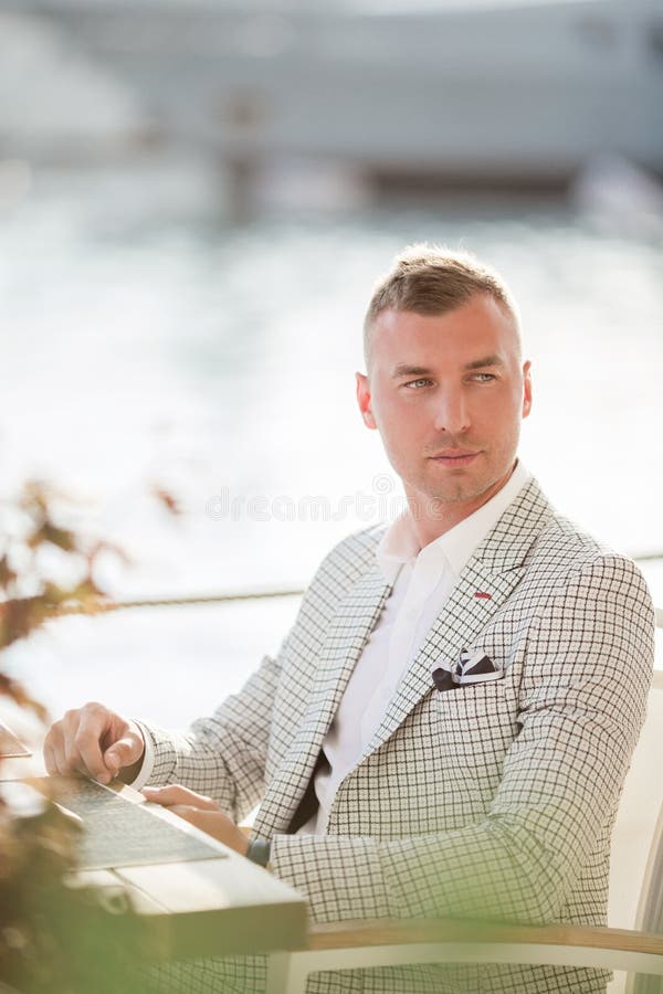 El hombre de negocios hermoso está llevando el traje blanco de la moda que se sienta en café con el fondo del mar Bah?a de Monten fotos de archivo libres de regalías El hombre de negocios hermoso está llevando el traje blanco de la moda que se sienta en café con el fondo del mar Bah?a de Monten fotos de archivo libres de regalías