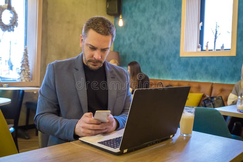 El hombre de negocios hermoso se está sentando en café moderno con el teléfono y el ordenador portátil elegantes y está gozando d imágenes de archivo libres de regalías El hombre de negocios hermoso se está sentando en café moderno con el teléfono y el ordenador portátil elegantes y está gozando d imágenes de archivo libres de regalías