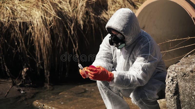 El hombre en traje del bio-peligro y careta antigás comprueba la contaminación del agua afuera