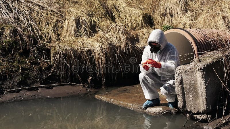 El hombre en traje del bio-peligro y careta antigás comprueba la contaminación del agua afuera