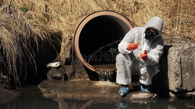 El hombre en traje del bio-peligro y careta antigás comprueba la contaminación del agua afuera