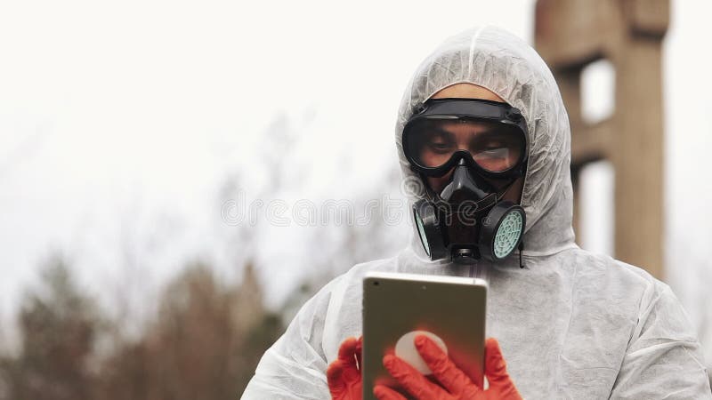 El hombre en traje del bio-peligro y careta antigás toma notas en su tableta que se coloca en la tierra contaminada