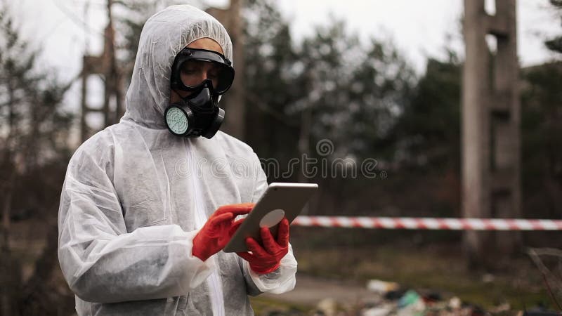 El hombre en traje del bio-peligro y careta antigás toma notas en su tableta que se coloca en la tierra contaminada