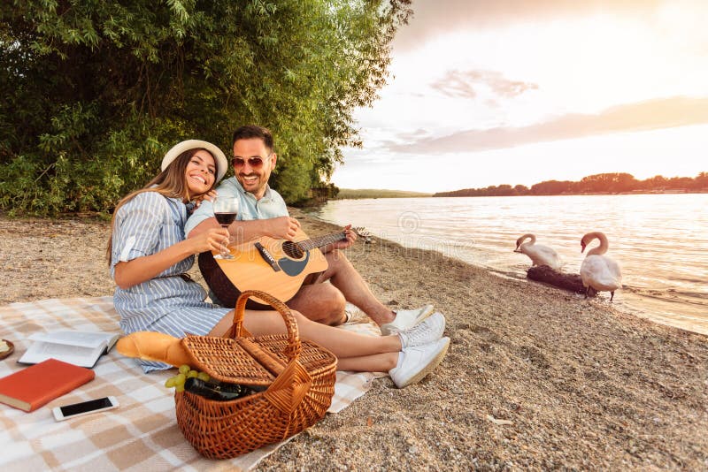 El hombre está tocando la guitarra y su novia está descansando su cabeza en su hombro Puesta del sol sobre el agua en el fondo foto de archivo libre de regalías El hombre está tocando la guitarra y su novia está descansando su cabeza en su hombro Puesta del sol sobre el agua en el fondo foto de archivo libre de regalías