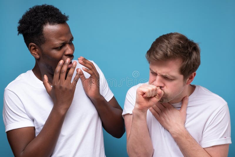 One man is coughing because of flu or covid. Problem with lungs. Another is shocked that his friend is ill Studio shot on blue wall. One man is coughing because of flu or covid. Problem with lungs. Another is shocked that his friend is ill Studio shot on blue wall