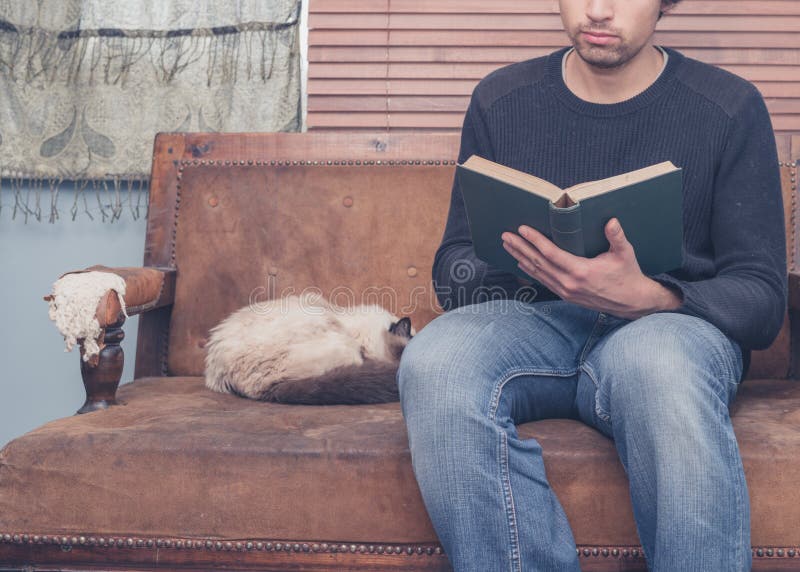 El hombre joven que se sienta en el sofá con el gato está leyendo fotos de archivo libres de regalías El hombre joven que se sienta en el sofá con el gato está leyendo fotos de archivo libres de regalías