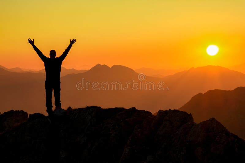 El hombre que gana del éxito feliz arma para arriba en la montaña en la puesta del sol