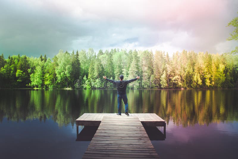 El hombre se coloca en el embarcadero de un lago hermoso, hombre joven que disfruta de vida, sus brazos se abre, un arco iris sob fotos de archivo libres de regalías