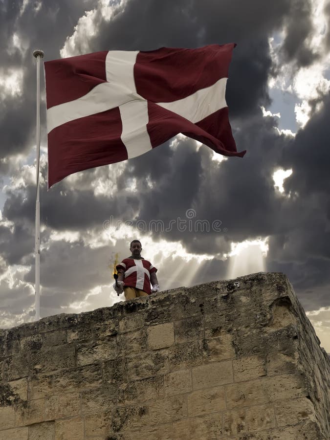 VITTORIOSA, MALTA - APR 24 - A Knights of the Order of St. John stands beneath the flag during filming of Valletta Living History. VITTORIOSA, MALTA - APR 24 - A Knights of the Order of St. John stands beneath the flag during filming of Valletta Living History