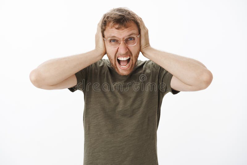 Guy lose temper being with loud noises at night yelling out loud with angry outrage expression covering ears with palms on head being in rage over white background in casual dark-green t-shirt. Guy lose temper being with loud noises at night yelling out loud with angry outrage expression covering ears with palms on head being in rage over white background in casual dark-green t-shirt.