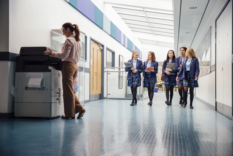 Group of teenage girls are walking down the school hall with books and laptops in their arms. They are talking and laughing as they walk and there is a female teacher using the printer. Group of teenage girls are walking down the school hall with books and laptops in their arms. They are talking and laughing as they walk and there is a female teacher using the printer.